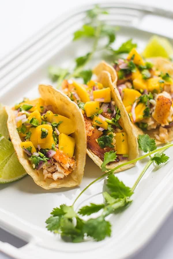 Three assembled bass fish tacos with mango salsa, cilantro, and lime wedges on a white tray